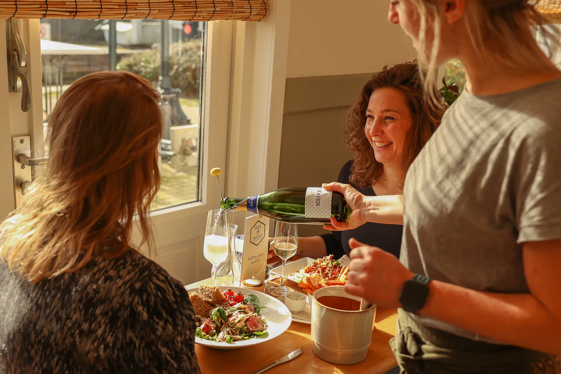 Medewerker schenkt bubbels in voor twee lachende gasten aan een tafel bij het raam