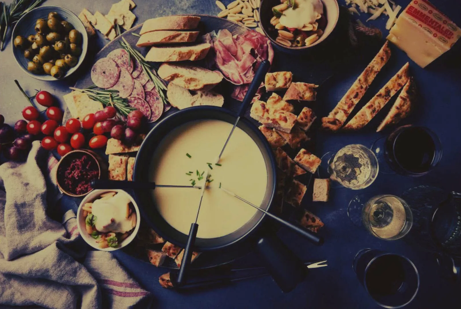 Bovenaanzicht van een tafel met kaasfondue, stokbrood, charcuterie, olijven, tomaatjes en glazen wijn