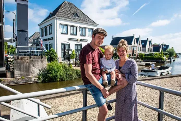 Mark en Stef van Veen met een peuter op een brug aan het water, met huizen en een sloep op de achtergrond