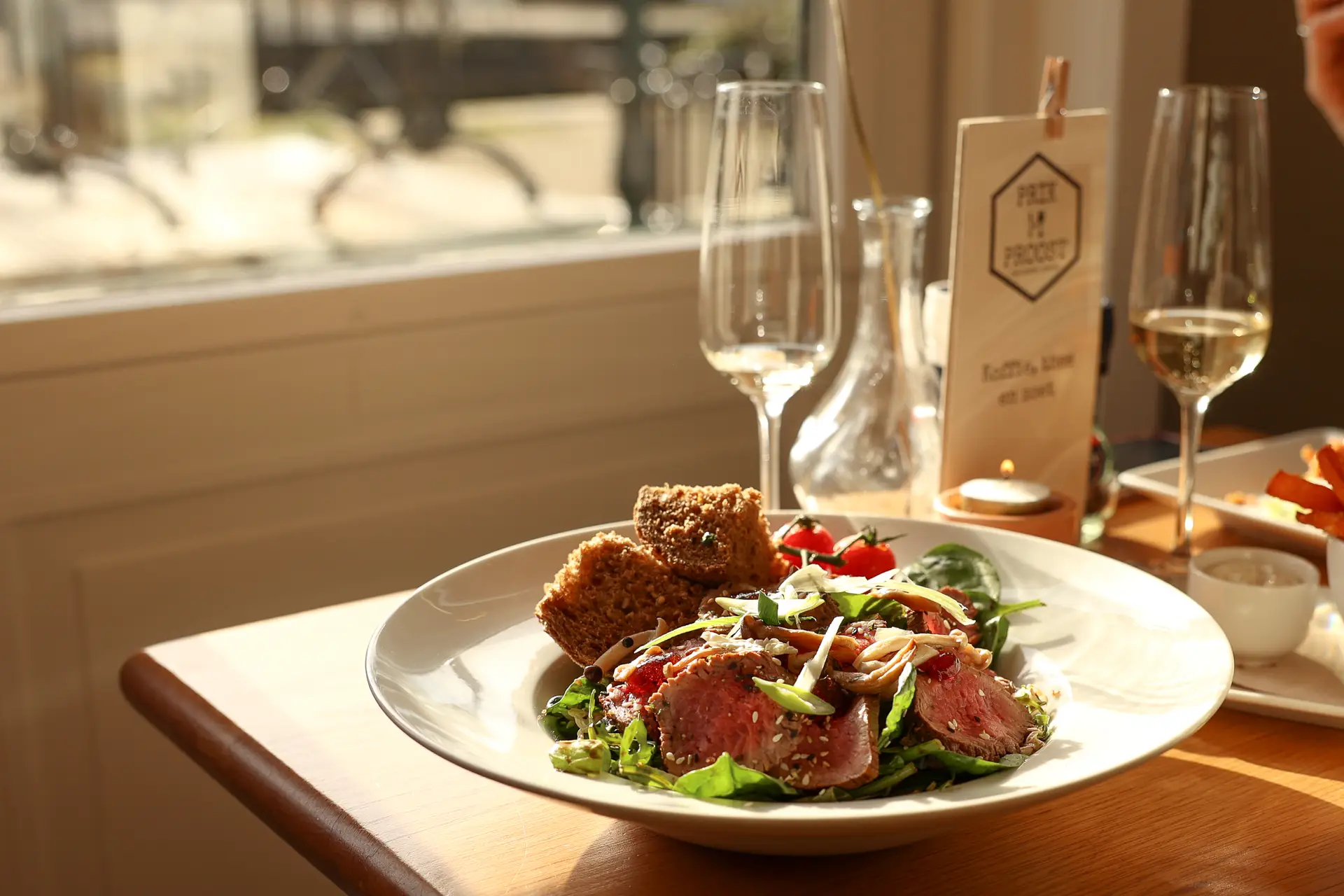 Salade met dun gesneden rosé vlees en geroosterd brood, met glazen witte wijn op tafel